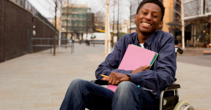 African American student in a wheelchair smiling at the camera while holding a pencil and notepads.