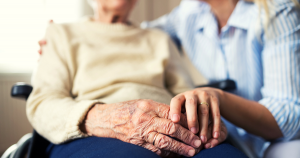 An elderly woman, sitting in a wheelchair, holding hands with another woman kneeling down beside her.