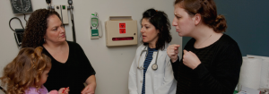 A small girl in doctor's office on exam table next to mother with doctor and interpreter.