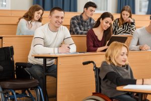Cheerful student looking at camera, smiling and posing while making notes during lecture. Students with wheelchairs receiving education and enjoying studying process.