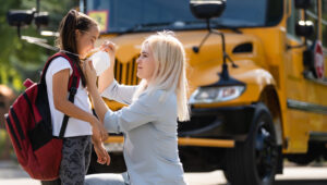 Caucasian woman puts a protective mask on her daughter outdoors. Caring mother helps to wear a mask schoolgirl near the school bus. Quarantine during coronavirus.