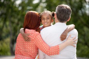 Hugging foster parents. Dark-skinned beaming girl feeling happy while hugging her foster parents