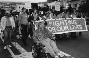 Image of dozens of protestors, including a person in a wheelchair, holding a sign that states "Fighting for our lives."