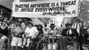 Image of participants in one of the first Disability Independence Day marches in New York City gathered under a banner that reads “‘Injustice Anywhere is a Threat to Justice Everywhere,’—Martin Luther King, Jr.”