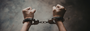 Close-up of a person’s wrists in handcuffs, with clenched fists raised against a blurred background.