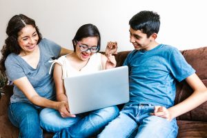 A woman and two children sit together on a couch smiling and looking at a laptop computer. The child in the center wears glasses and appears excited while the others look on with joy and engagement.