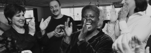 A black-and-white photograph shows disability rights advocate Lois Curtis at the center of a group of supporters. Curtis is smiling broadly and clapping as people around her applaud and celebrate. Several individuals stand nearby with their hands raised in applause, creating a joyful and triumphant atmosphere. The image captures a moment of celebration associated with the landmark Olmstead v. L.C. decision, which helped advance the rights of people with disabilities to live in their communities rather than be unnecessarily institutionalized.